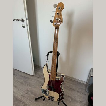Elektrická basová kytara v krémové barvě s tmavě červeným pickguardem, umístěná na černém stojanu. Na hlavě nástroje je nápis „EMELIA PRECISION BASS“…