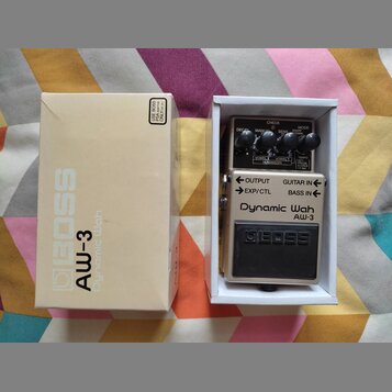Krabice a efekt pedál značky BOSS model AW-3 Dynamic Wah. Pedál je umístěn v bílé krabičce a má stříbrné tělo s černým spínacím pedálem, na kterém je…
