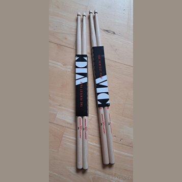 Dvě dvojice dřevěných bubeníckých paliček v původním papírovém obalu. Na paličkách je červený nápis AMERICAN CLASSIC 7A a logo značky VIC FIRTH. Obal…