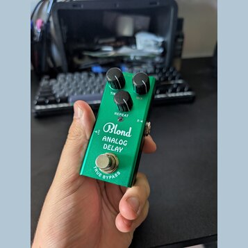 Zelený efekt pedál s kovovým povrchem. Na přední straně je bílý nápis „Blond ANALOG DELAY“ a v dolní části text „TRUE BYPASS“. Pedál má tři černé ovl…