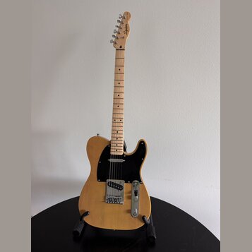 Elektrická kytara typu Telecaster v přírodní dřevěné barvě s černým pickguardem. Na hlavě hmatníku je vidět nápis Squier. Nástroj je umístěn na černé…
