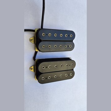Dvě černé elektrické kytarové snímače typu humbucker s kovovými zlatými rámečky. Každý snímač má dvě řady kulatých pólů. Povrch plastových krytů je h…