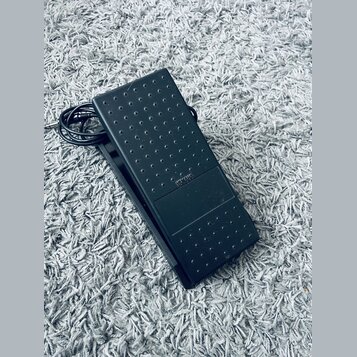 Černý sustain pedál z plastu s texturovaným povrchem v podobě vyvýšených teček. Uprostřed je viditelný nápis YAMAHA. K pedálu vede černý kabel s kone…