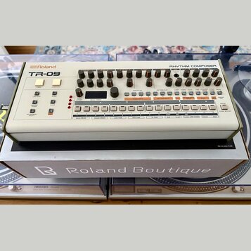 Bílá rytmická sekvenční jednotka s nápisy Roland a TR-09 v horní levé části a RHYTHM COMPOSER v pravé. Zařízení obsahuje řadu černých otočných ovlada…