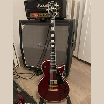 Červená elektrická kytara typu Les Paul s bílým bindingem a zlatými potenciometry a hardwarem. Na hlavě hmatníku je nápis Gibson a logo, na hmatníku…
