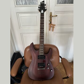Elektrická kytara s tmavě hnědým, polomatným povrchem těla a hnedým hmatníkem. Na hlavě je bílý nápis Schecter Diamond Series. Nástroj má dva humbuck…