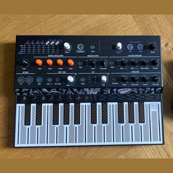 Černý kompaktní syntetizátor s nápisem MICROFREAK SYNTHESIZER. Horní panel obsahuje různé ovládací prvky, knoflíky a nápisy jako DIGITAL OSCILLATOR,…