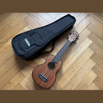 Hnědý dřevěný ukulele s černým hmatníkem a černým mostkem. Na hlavě nástroje je bílý nápis CASOCHA. Vedle nástroje leží černý textilní obal s bílým n…