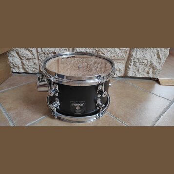 Černý buben s chromovaným obručováním a mechanismy. Na korpusu je nalepený štítek s nápisy SONOR, Sonic Rain a MADE IN GERMANY. Na horním blaně je vi…
