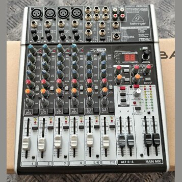 Stříbrný mixážní pult značky behringer s černým šasi. Na horním panelu je nápis 2-TRACK/USB a modelové označení XENYX X1204USB. Zařízení obsahuje něk…