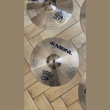 Fotografie zobrazuje dva kovové činely v tradičním zlatavě hnědém odstínu s viditelným rýhováním. Na spodním činele je nápis MEINL a logo značky, dál…