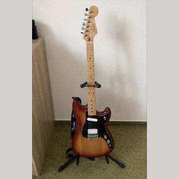 Elektrická kytara s tělem v červenohnědém sunburst provedení a světlým javorovým hmatníkem. Na hlavě je viditelné logo Fender a na popruhu nápis Fend…