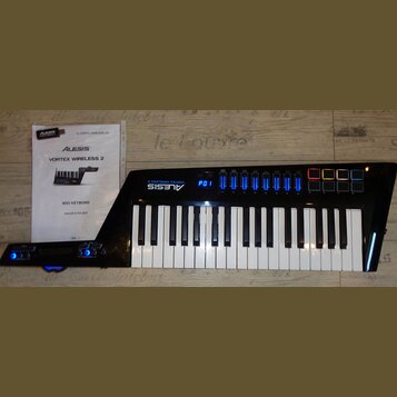 Černý MIDI kontrolér s klávesnicí a modrým podsvícením displeje a indikátorů. Na přístroji je bílý nápis ALESIS VORTEX WIRELESS 2. Vedle nástroje lež…