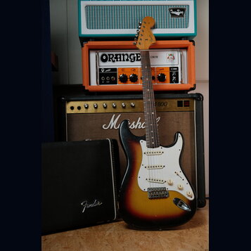 Elektrická kytara typu Stratocaster s barevným přechodem od žluté přes oranžovou k hnědé a bílým pickguardem. Na hlavě krku je viditelné logo Fender.…