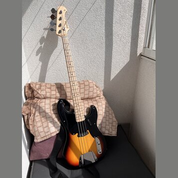 Elektrická basová kytara s tělem v barevném přechodu od žluté přes oranžovou k červené (sunburst) a černým pickguardem. Na hlavě hmatníku je vidět ná…