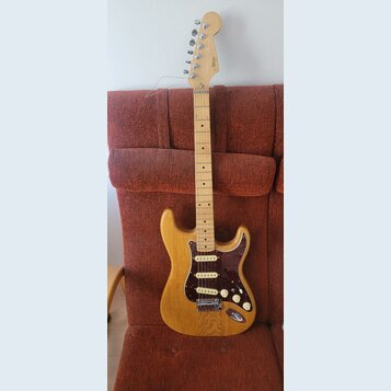 Elektrická kytara typu Stratocaster s přírodním dřevěným povrchem těla a hmatníkem. Tělo má tmavě červený želvový pickguard a tři bílé snímače. Na hl…