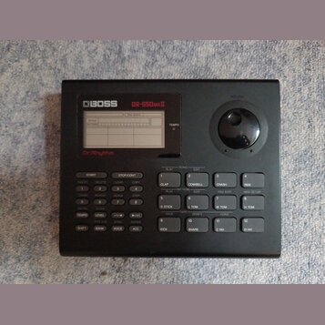 Černý rytmický automat s nápisy BOSS a DR-550MKII. Pod displejem je červený nápis Dr.Rhythm. Zařízení obsahuje řadu ovládacích tlačítek s různými pop…