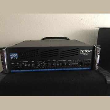 Černý basový zesilovač s kovovým šasi a madlem. Na předním panelu je vlevo logo EBS a vpravo nápis TD650 Professional Tube Definition Bass Head. Dole…