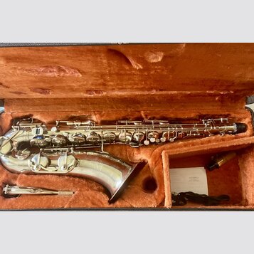 Stříbrný saxofon v otevřeném kufru s oranžovou plyšovou výstělkou. Nástroj má lesklý kovový povrch a je vybaven kompletní mechanikou s perleťovými kl…