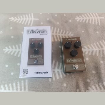 Na fotografii je efekt pedál Analog Delay značky tc electronic, model Echobrain, v hnědé barvě s kovovým šasi. Pedál má tři černé ovládací knoflíky s…