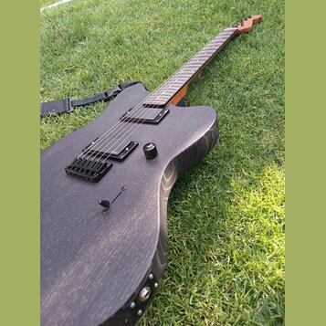 Černá elektrická kytara s matným povrchem a viditnou strukturou dřeva, položená na trávě. Nástroj má dva humbuckery, jeden ovládací knoflík a černý m…