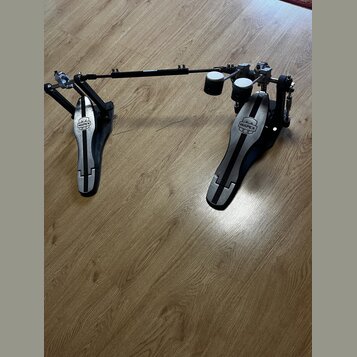 Dvojpedál na bicí v černé a stříbrné barvě s kovovými komponenty. Na obou šlapkách je umístěno logo MAPEX. Povrch pedálů vykazuje známky běžného použ…