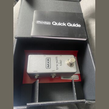 Stříbrný efekt pedál značky MXR v černé krabici, vedle kterého leží černý manuál s nápisem Dunlop Quick Guide. Na pedálu jsou nápisy MXR, trs split +…