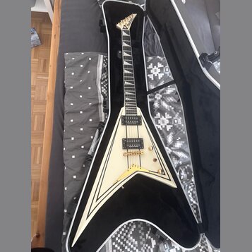 Bílá elektrická kytara s tvarem V v černém obalu. Na hlavě hmatníku je nápis Jackson. Kytara má dva humbuckery, zlatý hardware a bílý lem kolem těla.…