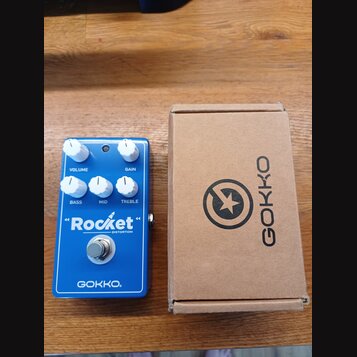 Modrý efekt pedál s nápisy "Rocket" DISTORTION a GOKKO. Na horní straně jsou čtyři bílé ovládací knoflíky s popisky VOLUME, GAIN, BASS, MID a TREBLE.…