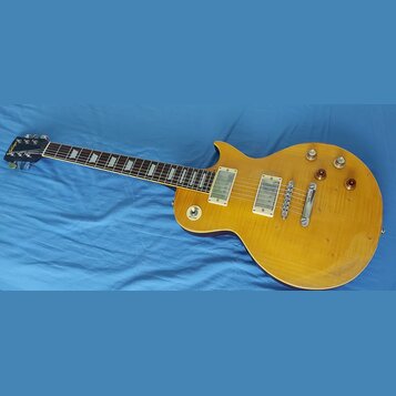 Žlutá elektrická kytara typu Les Paul s hmatníkem z tmavého dřeva a trapezoidními vložkami. Na hlavě je nápis Gibson. Kytara má dva humbuckery s kryt…