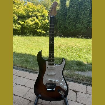 Elektrická kytara s trupem v tmavě hnědé barvě s přechodem do černé a bílým pickguardem. Na hlavě hmatníku je viditelný nápis Fender. Nástroj má tři…