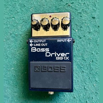 Modrý efekt pedál s nápisy Bass Driver a BB-1X. Horní část je z anodizovaného kovu se čtyřmi ovládacími knoflíky, červenou kontrolkou a nápisem CHECK…