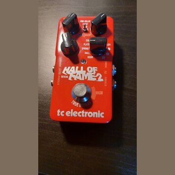Červený efekt pedál s nápisem HALL OF FAME 2 REVERB a logem tc electronic. Na horní části jsou tři černé otočné ovladače a přepínače s popisky PRE-DE…