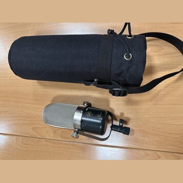 Stříbrný kondenzátorový mikrofon s černým tělem a kovovým držákem. Na těle je viditelné logo značky AKG a nápis R1 ACM-180. Vedle mikrofonu je černý…