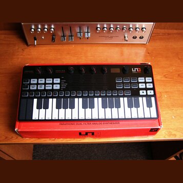 Černý analogový syntetizátor s klaviaturou, umístěný v červeném obalu. Na horním panelu jsou nápisy ANALOG SYNTHESIZER, PARAPHONIC DUAL FILTER a logo…