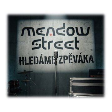 Fotografie zachycuje stříbrný mikrofon na stojanu umístěný před šedou stěnou. Na stěně je černý nápis „mendow street HLEDÁME ZPĚVÁKA“. V pozadí jsou…