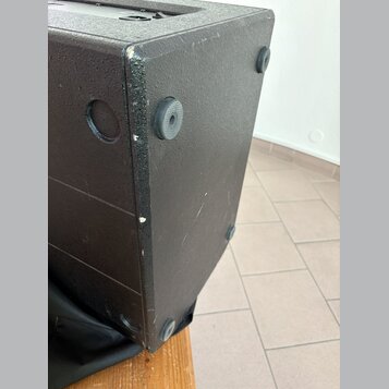 Detail černé reproduktorové bedny s texturovaným povrchem. Na spodní straně jsou vidět čtyři gumové nožičky a na boku kulatý úchop. Hrany bedny vykaz…