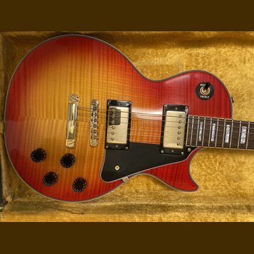 Elektrická kytara typu Les Paul v červeno-žlutém sunburst provedení s viditnou strukturou dřeva. Nástroj má dva snímače s krémovými kryty, zlatý most…