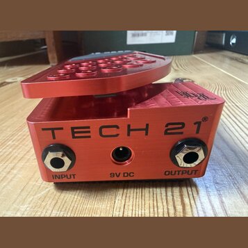 Červený kytarový efekt pedál z anodizovaného hliníku. Na přední straně je velký černý nápis TECH 21® a označení vstupů INPUT, 9V DC a OUTPUT. Povrch…
