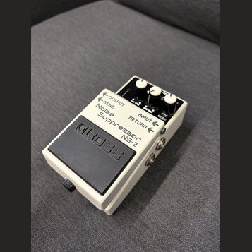 Bílý efekt pedál Boss Noise Suppressor NS-2 v kompaktním kovovém provedení s černým gumovým spínacím pedálem. Na horní části jsou čtyři bílé ovládací…