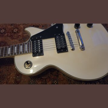 Bílá elektrická kytara s černým pickguardem a dvěma černými humbuckery. Na přepínači tónu jsou viditelné nápisy TREBLE a RHYTHM. Nástroj má chromovan…