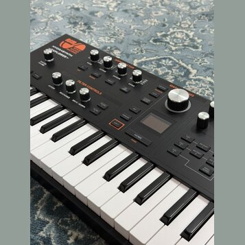 Černý klávesový syntetizátor s bílými a černými klávesami. Na horním panelu jsou nápisy ASM, SONIC MACHINES a model HYDRASYNTH EXPLORER. Přístroj obs…