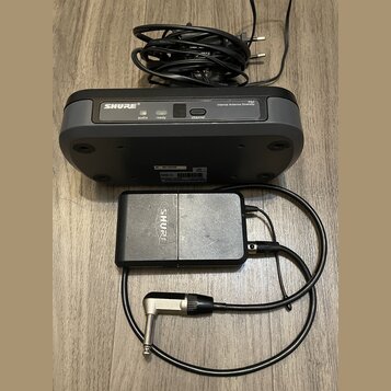 Černý bezdrátový vysílač a přijímač značky SHURE. Na horním zařízení je nápis SHURE, model PG4 a popis Internal Antenna Diversity s kontrolkami audio…