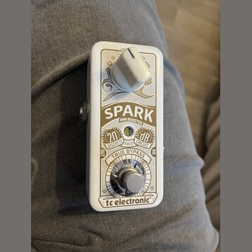Bílý efekt pedál Spark Mini Booster značky tc electronic. Na přední straně je bílý oválný ovládací knoflík a stomečový spínač True Bypass. Jsou zde n…