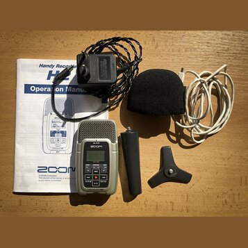 Sada obsahuje stříbrný digitální záznamník s nápisy ZOOM, MIC ACTIVE a Handy Recorder H2. K zařízení patří černý napájecí adaptér s nápisem CAUTION A…