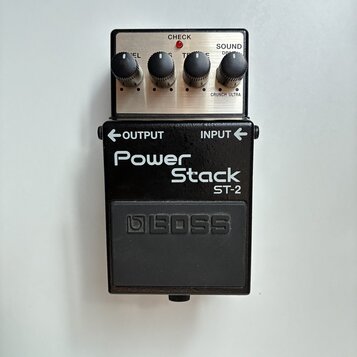 Kytarový efekt pedál značky BOSS, model Power Stack ST-2. Zařízení má černý kovový korpus s nápisy OUTPUT a INPUT u konektorů. Horní část tvoří stříb…