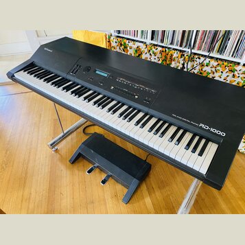 Černý digitální klavír na kovovém stojanu s přiloženým černým pedálem. Na horní části je nápis Roland a vpravo u kláves označení ROLAND DIGITAL PIANO…
