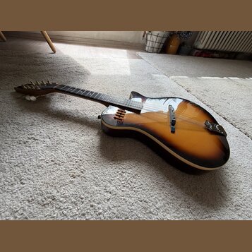 Akustická kytara s korpusem v přechodovém žlutě-hnědém odstínu (sunburst) a tmavým hmatníkem. Nástroj leží na světlém koberci, má bílé ladičky a kový…
