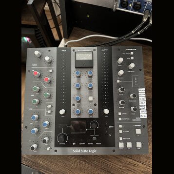 Černý rackový procesor s nápisem Solid State Logic. Zařízení obsahuje analogové VU metr, několik bílých a modrých otočných knoflíků, posuvníky a bílé…