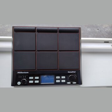 Černý elektronický bicí pad s devíti čtvercovými polštáři v mřížce 3x3. Na spodním panelu jsou nápisy Millenium a NonaPad, malý displej, několik tlač…